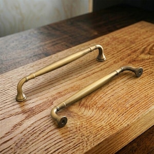 Könnte beinhalten: Zwei antike Messing-Schrankgriffe mit gebogenen Griffen und Montagematerial. Die Griffe werden auf einer Holzoberfläche präsentiert, die ihr Vintage-Design und ihre Oberfläche zeigt. Ideal für Restaurierungsprojekte.