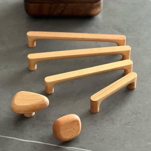 May include: A selection of wooden cabinet hardware, featuring four rectangular handles and two rounded knobs. The light-coloured wood hardware is arranged on a grey surface. The handles are of varying sizes, suitable for different cabinet dimensions.