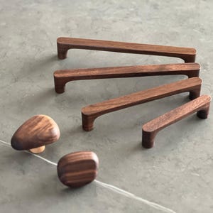 May include: A selection of wooden cabinet hardware, featuring four pull handles and two knobs. The handles are rectangular with a curved grip, varying in size. The knobs are rounded and mushroom-shaped. All items are crafted from dark brown wood.