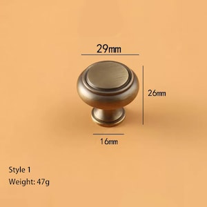 May include: Bronze-toned cabinet knob, Style 1, with a diameter of 29mm, a height of 26mm, and a base of 16mm. The knob weighs 47g. The design features a rounded top and a decorative base.