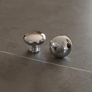 以下が含まれることがあります： 2つの光沢のあるクロムキャビネットノブ。1つは短いステムが付いた丸みを帯びた楕円形で、もう1つは球体です。どちらのノブも周囲の環境を反射しています。ノブは灰色の表面にあります。