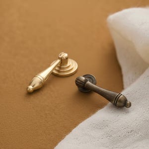 Może przedstawiać: Dwa uchwyty do szuflad w stylu antycznym. Jeden jest w kolorze złotym z zaokrągloną podstawą i uchwytem w kształcie łzy. Drugi jest w kolorze brązu z okrągłą podstawą i żebrowanym uchwytem. Oba leżą na brązowej powierzchni.