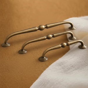 Może przedstawiać: Trzy uchwyty do szafek z antycznego mosiądzu w różnych rozmiarach. Każdy uchwyt ma zakrzywiony uchwyt z centralną kulą dekoracyjną i zaokrąglonymi płytkami końcowymi. Uchwyty są ułożone na teksturowanej brązowej powierzchni z białym materiałem w tle.