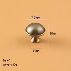 May include: Antique brass-finished cabinet knob, Style 2, with a rounded top and a pedestal base. The knob measures 29mm wide, 28mm high, and 14mm deep. It weighs 42g. The knob is designed for furniture hardware.