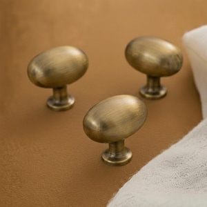 May include: Three oval-shaped cabinet knobs with a brushed antique brass finish. Each knob features a smooth, rounded top surface and a cylindrical base that connects to a circular mounting plate, showcasing a metallic texture and warm gold tones.