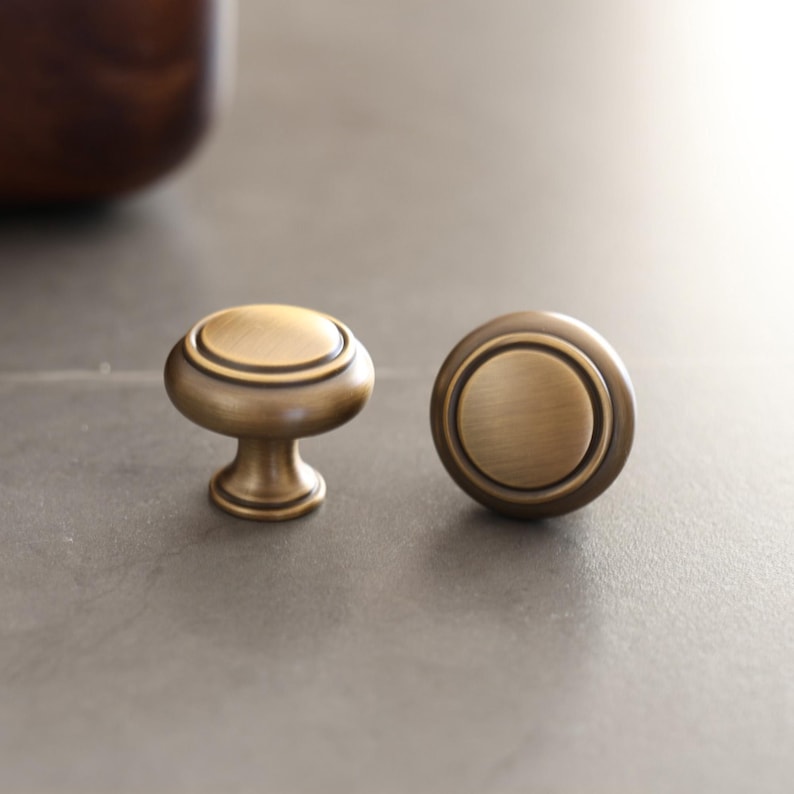 May include: Two antique brass cabinet knobs. One knob is mounted on a small pedestal, while the other is a flat disc shape. Both knobs have a circular design with a raised outer ring. The knobs are made of metal and have a warm, golden color.