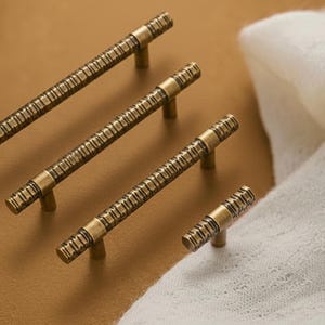 Könnte beinhalten: Ein Satz von vier antiken Messing-Schrankgriffen in verschiedenen Größen. Die Griffe haben ein strukturiertes, lineares Design und eine warme, gealterte Oberfläche. Die Hardware ist auf einem neutralen Hintergrund angeordnet.