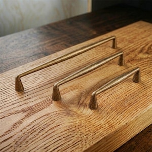 May include: Three bronze-colored cabinet pulls of varying sizes are arranged on a wooden surface. The pulls have a simple, elongated design with slightly curved handles and angled feet. The wood grain is visible.