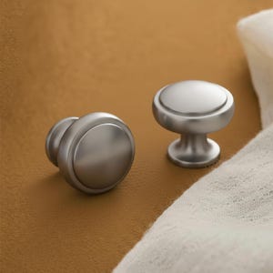 May include: Two brushed nickel cabinet knobs. One knob is on its side, and the other is upright. The knobs have a rounded top and a cylindrical base. The background is a warm, neutral colour.