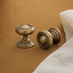May include: Two round cabinet knobs made of antique brass with a brushed finish. Each knob features a smooth, domed centre surrounded by a decorative rope-style border and a sturdy, tapered base for mounting to furniture or cabinetry doors.