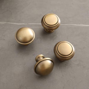 May include: Four antique brass cabinet knobs with a rounded design. The knobs have a circular top and a decorative base. The finish is a warm, aged brass color, and they are arranged on a gray surface.