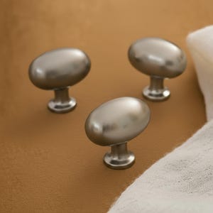 Könnte beinhalten: Drei ovale Schrankknöpfe aus gebürstetem Nickel. Die Knöpfe haben eine glatte, ovale Form und sind auf einer kurzen, zylindrischen Basis montiert. Die Hardware befindet sich auf einer braunen Oberfläche mit einem weißen Tuch im Hintergrund.