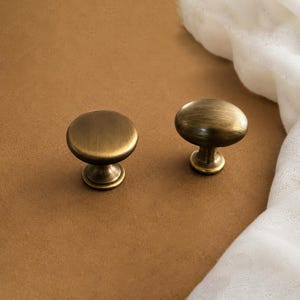 May include: Two round cabinet knobs with a brushed antique brass finish. Each knob features a smooth, slightly domed top surface and a tapered base, showcasing a metallic texture with subtle dark highlights across the circular hardware pieces.