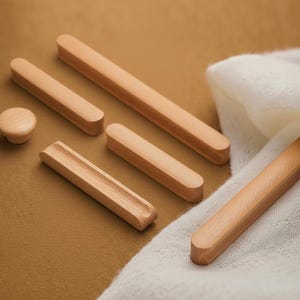 Könnte beinhalten: Eine Sammlung unfertiger Holzmöbelbeschläge, darunter ein runder Knopf, mehrere rechteckige Griffe und ein gerillter Griff. Die Teile sind aus hellem Holz gefertigt und auf einer braunen Oberfläche mit einem weißen Tuch im Hintergrund angeordnet.