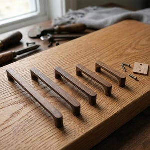 Op de afbeelding: Een verzameling houten kastgrepen in verschillende maten, gerangschikt op een houten oppervlak. De grepen zijn diep bruin van kleur, met een rechthoekig ontwerp en afgeronde randen. Schroeven en een klein houten sjabloon zijn ook aanwezig.
