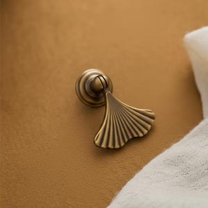 May include: A brass-toned metal cabinet pull featuring a circular base and a hinged, fan-shaped handle with vertical ridges. The hardware has a dark, antiqued finish with highlights on the raised edges, creating a distinct contrast in color.