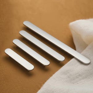 May include: Four brushed nickel cabinet pulls of varying lengths are displayed. The pulls have a flat, rectangular design with rounded ends. They are set against a tan backdrop, with a white cloth in the background.