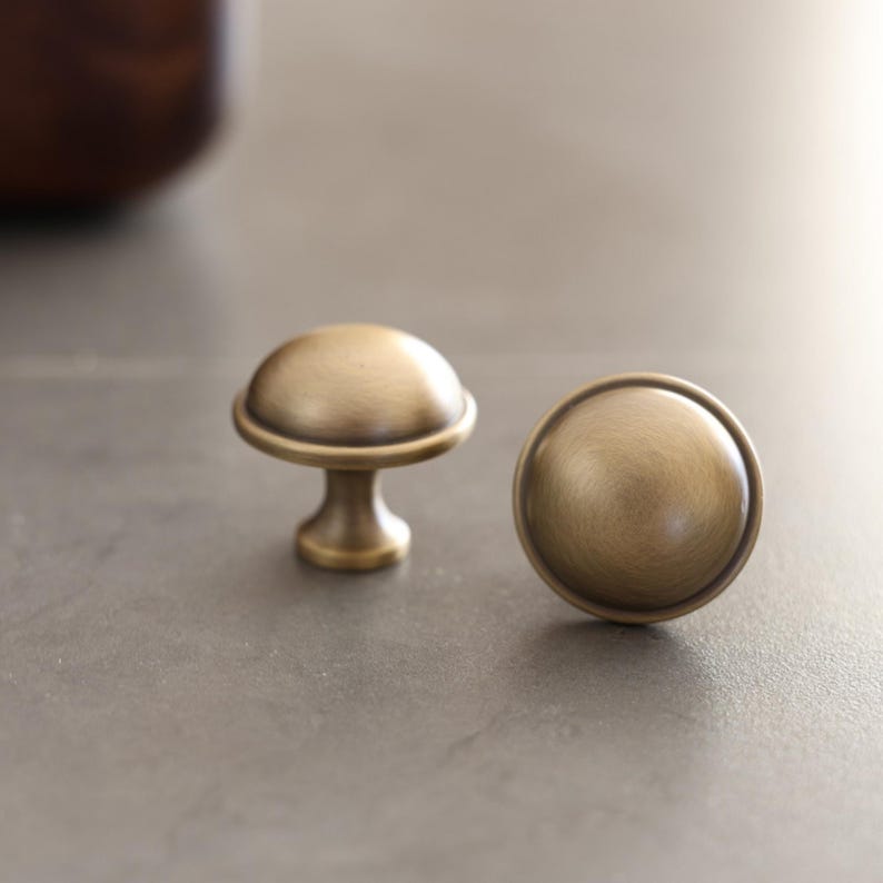 May include: Two antique brass cabinet knobs. The knobs have a rounded, mushroom-like shape with a circular base and a domed top. The brass has a warm, aged patina, suggesting vintage or rustic decor.