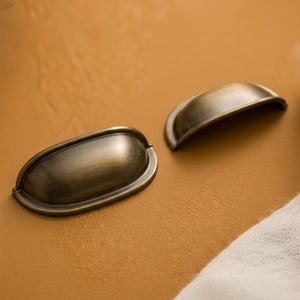 May include: Two antique bronze-finished drawer pulls. The pulls have a half-moon shape with a rounded, oval base. The metal has a distressed finish, giving it a vintage appearance. These pulls are ideal for adding a classic touch to furniture.