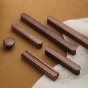 May include: A collection of wooden cabinet hardware, including knobs and various sizes of rectangular pulls. The hardware is made of dark brown wood and is arranged on a tan surface with a white cloth.