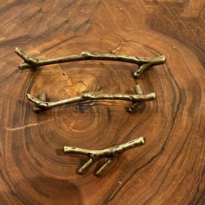 May include: Three antique gold-toned metal cabinet pulls shaped like branches. The pulls are arranged in a row on a wooden surface.