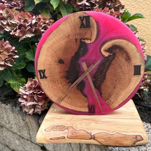Könnte beinhalten: Eine runde Holzuhr mit einem pinkfarbenen Harzmittelteil und römischen Ziffern. Das Zifferblatt weist natürliche Holzmaserungsmuster auf und ist auf einem Holzständer montiert. Die Uhrzeiger sind aus Holz.