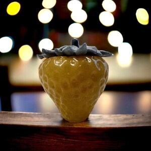 May include: A yellow ceramic strawberry-shaped container with a gray lid. The container is sitting on a wooden surface.