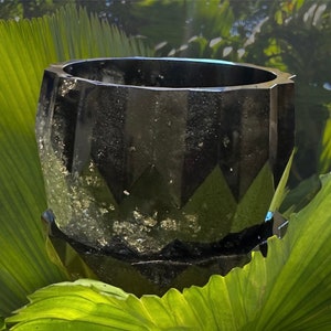 May include: A black, faceted, geometric planter with a shiny, almost metallic finish. The planter is sitting on a bed of green leaves.