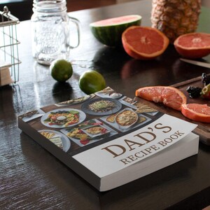 May include: A white cookbook with the title "Dad's Recipe Book" on a black table. The cookbook is open to a page with photos of various food dishes. There are also various fruits and vegetables on the table, including a pineapple, watermelon, grapefruit, and limes.