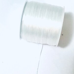 May include: A spool of clear, elastic beading cord. The cord is tightly wound on a white plastic spool. A length of the cord extends from the spool, suggesting it is ready for use in jewelry making or crafts.