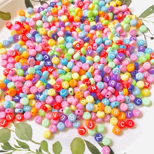 May include: A large pile of colorful plastic alphabet beads. The beads are in various pastel colors, including pink, blue, yellow, green, and orange. Each bead has a white letter printed on it.