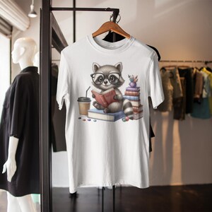 A white t-shirt with a graphic design of a raccoon wearing glasses and holding a book, surrounded by books and other items.