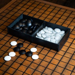 May include: A Go game set featuring black and white stones on a wooden board. The stones are kept in a black leather tray with two sections. The board has a grid pattern, and some stones are scattered around the tray.