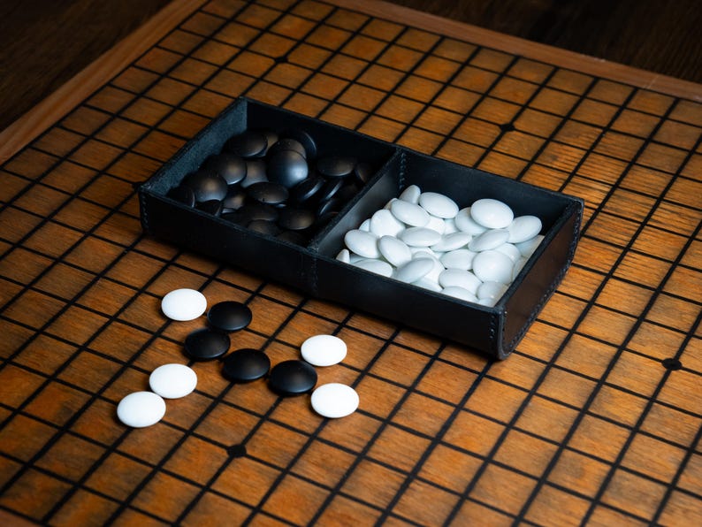 May include: A Go game set featuring black and white stones on a wooden board. The stones are kept in a black leather tray with two sections. The board has a grid pattern, and some stones are scattered around the tray.