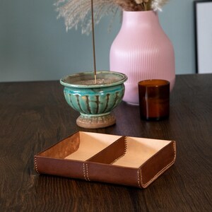 May include: A brown leather tray with two compartments, stitched with white thread. The tray sits on a dark wooden surface, with a teal ceramic bowl, a pink vase, and a brown glass container in the background.