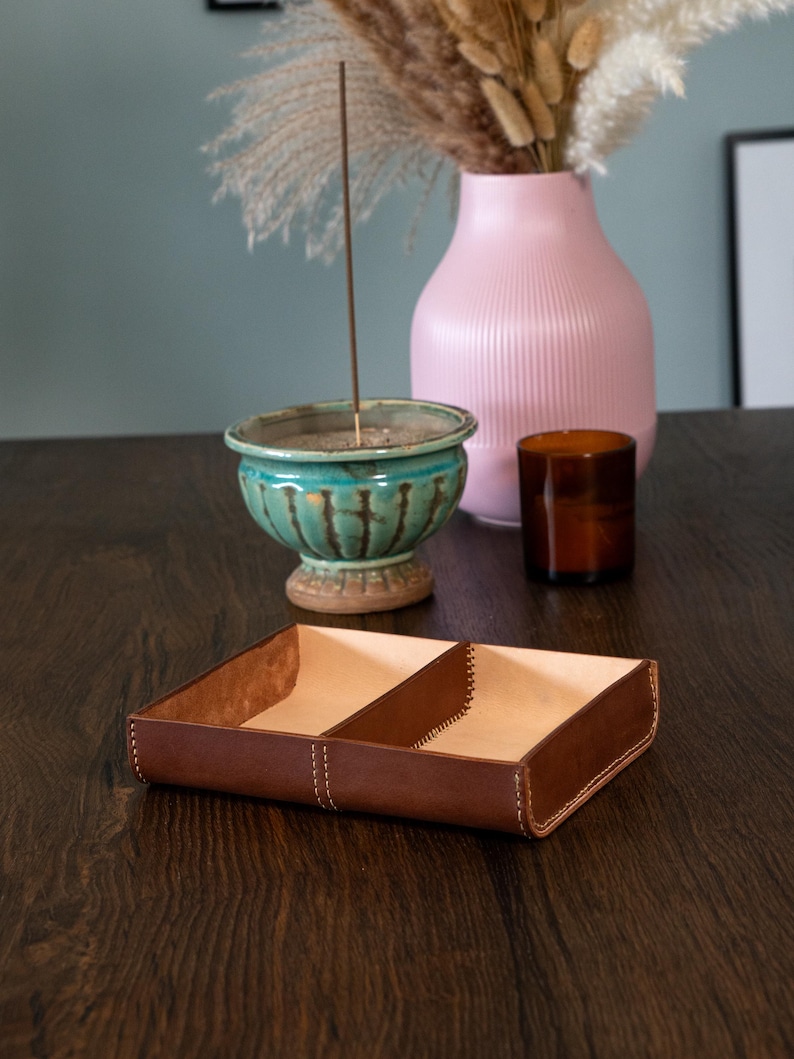 May include: A brown leather tray with two compartments, stitched with white thread. The tray sits on a dark wooden surface, with a teal ceramic bowl, a pink vase, and a brown glass container in the background.