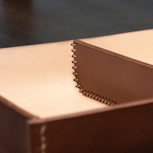 May include: A close-up shot of a brown leather box with a light tan interior. The box is divided into sections and has visible stitching along the edges. The leather has a smooth texture, and the stitching is a contrasting light colour.