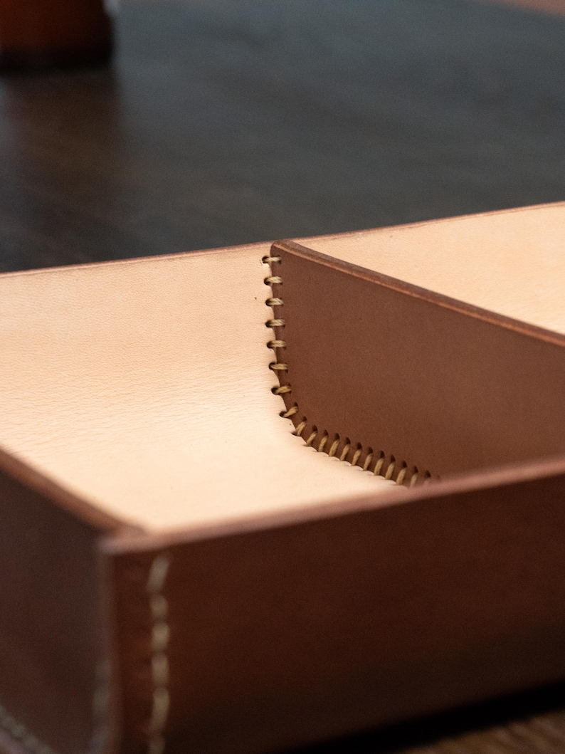 May include: A close-up shot of a brown leather box with a light tan interior. The box is divided into sections and has visible stitching along the edges. The leather has a smooth texture, and the stitching is a contrasting light colour.