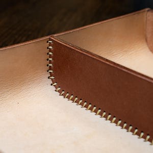 May include: Close-up of a handcrafted leather tray with a two-tone design. The exterior is a rich brown, while the interior has a lighter tan hue. The edges are meticulously stitched with a contrasting thread, showcasing the craftsmanship.