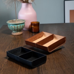 May include: Two leather trays, one brown and one black, each with three compartments, are on a wooden surface. The brown tray has a light-coloured interior. A pink vase, a teal bowl, and a small brown container are in the background.