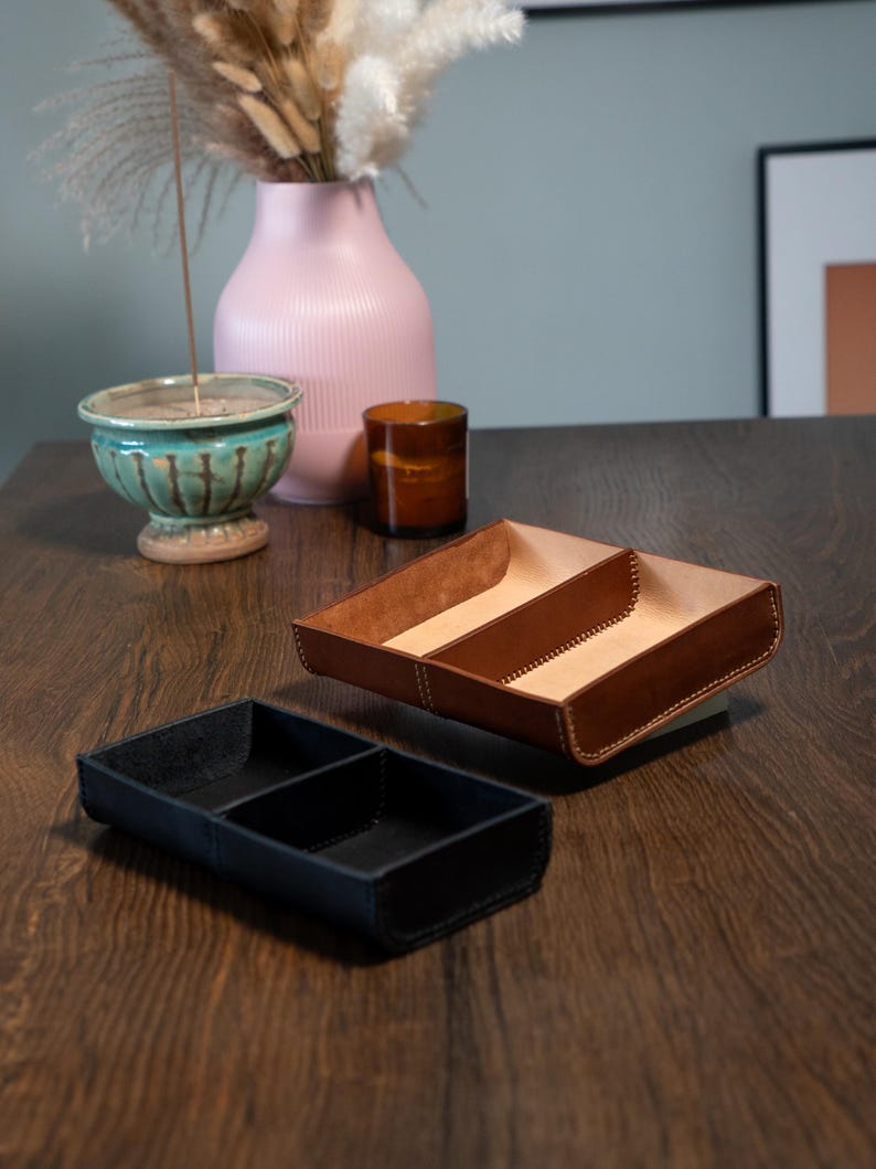 May include: Two leather trays, one brown and one black, each with three compartments, are on a wooden surface. The brown tray has a light-coloured interior. A pink vase, a teal bowl, and a small brown container are in the background.