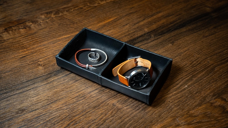 May include: A black leather tray with two compartments, containing a silver ring, a red and silver bracelet, and a wristwatch with a black face and tan leather strap. The tray is on a wooden surface.