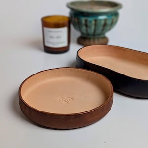 Könnte beinhalten: Zwei runde Ledertabletts, eines braun und eines schwarz, mit einem hellbraunen Innenraum. Das braune Tablett hat ein kleines geprägtes Design in der Mitte.