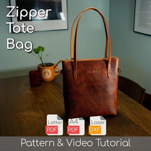 Op de afbeelding: Een bruine leren tote bag met lange handvatten, een ritssluiting en de tekst "Zipper Tote Bag". De tas staat op een houten tafel met een kleine plant en een rood glas. De afbeelding bevat ook PDF- en DXF-bestandspictogrammen.