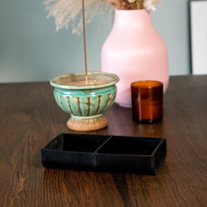 May include: A black leather tray with two sections on a dark wood surface. A teal ceramic incense holder with a lit incense stick, a pink vase with dried flowers, and a brown glass candle holder are in the background.