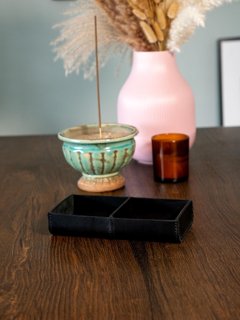 May include: A black leather tray with two sections on a dark wood surface. A teal ceramic incense holder with a lit incense stick, a pink vase with dried flowers, and a brown glass candle holder are in the background.