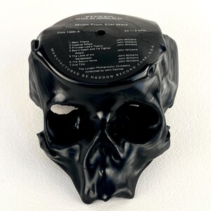 May include: A black skull sculpture made from a vinyl record. The record label is visible, with text that reads "Music From Star Wars" and a list of song titles. The record is molded into the shape of a skull.