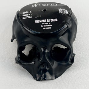 May include: A black skull-shaped object with a vinyl record on top. The record is black with white text that reads "RHOMBUS OF DOOM" and "MAGNETO Recordings". The skull has large eye sockets and a detailed nose and mouth.