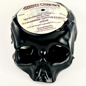 May include: A black skull sculpture made from a vinyl record. The record label is visible, with text including "HIGH PRIEST of HARMFUL MATTER" and song titles. The sculpture is on a white background.