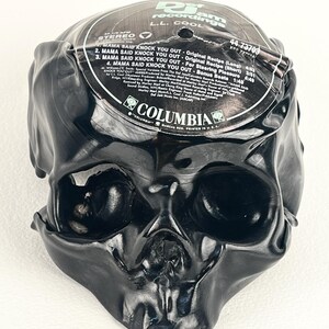 May include: A black skull sculpture made from a melted vinyl record. The record label is visible, with text including "MAMA SAID KNOCK YOU OUT" and "L.L. COOL". The sculpture has a glossy finish and detailed facial features.